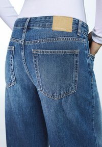 Modré džíny z denimu s ležérním střihem, vybavené dvěma zadními kapsami, prošitými detaily a hnědou koženou nášivkou na pásku.