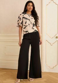 Kreemja ja musta lillemustriga pluus, millel on volditud ja volangiga varrukad, koos mustade laia jalgadega pükste ja valgete rihmadega kontsakingadega.