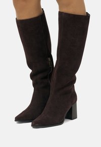 Stivaletti al ginocchio in suede marrone scuro con tacco a blocco e zip laterali indossati su gambe scoperte su uno sfondo bianco.