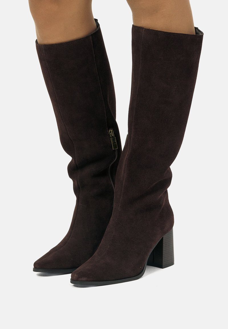 Stivaletti al ginocchio in suede marrone scuro con tacco a blocco e zip laterali indossati su gambe scoperte su uno sfondo bianco.