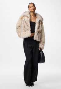 Kurtka ze sztucznego futra w beżowym kolorze z puszystym kołnierzem, ozdobiona cętkowanym wzorem. Noszona z czarnymi spodniami o szerokich nogawkach i teksturowaną torebką.