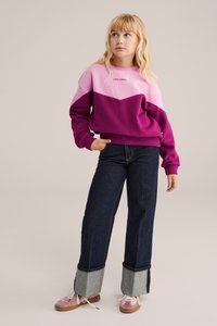 Roze un violetā krāsu blokā modei izgatavota džemperis ar uzrakstu "MĪLESTĪBA VISMŪŽ". Apvienots ar tumši ziliem platā kāju biksēm, ar pelēku denimu apakšējo malu. Rozā apavi.