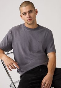 Γκρι κοντομάνικη μπλούζα από απαλό ύφασμα, με στρογγυλή λαιμόκοψη και διακριτικό ανάγλυφο λογότυπο στο στήθος. Το μοντέλο είναι καθιστό και φοράει μαύρο τζιν.