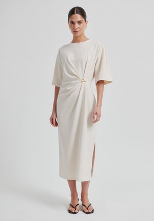 Beiges knielanges Kleid mit kurzen Ärmeln, ausgestattet mit einem gedrehten Detail an der Taille und einem goldenen Akzent. Glatter Stoff mit einem Seitenschlitz.