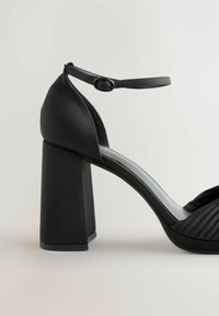 Sort højhælet sandal med bred blokhælhæl, justerbar ankelrem, glat satinmateriale og plisserede detaljer på tåen.