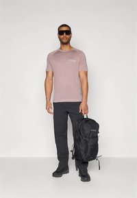 Hellrosa kurzärmliges T-Shirt mit rundem Halsausschnitt, schwarze Cargohose, schwarzer Rucksack und schwarze Schuhe; der Träger ist ein Modell, das aufrecht steht.