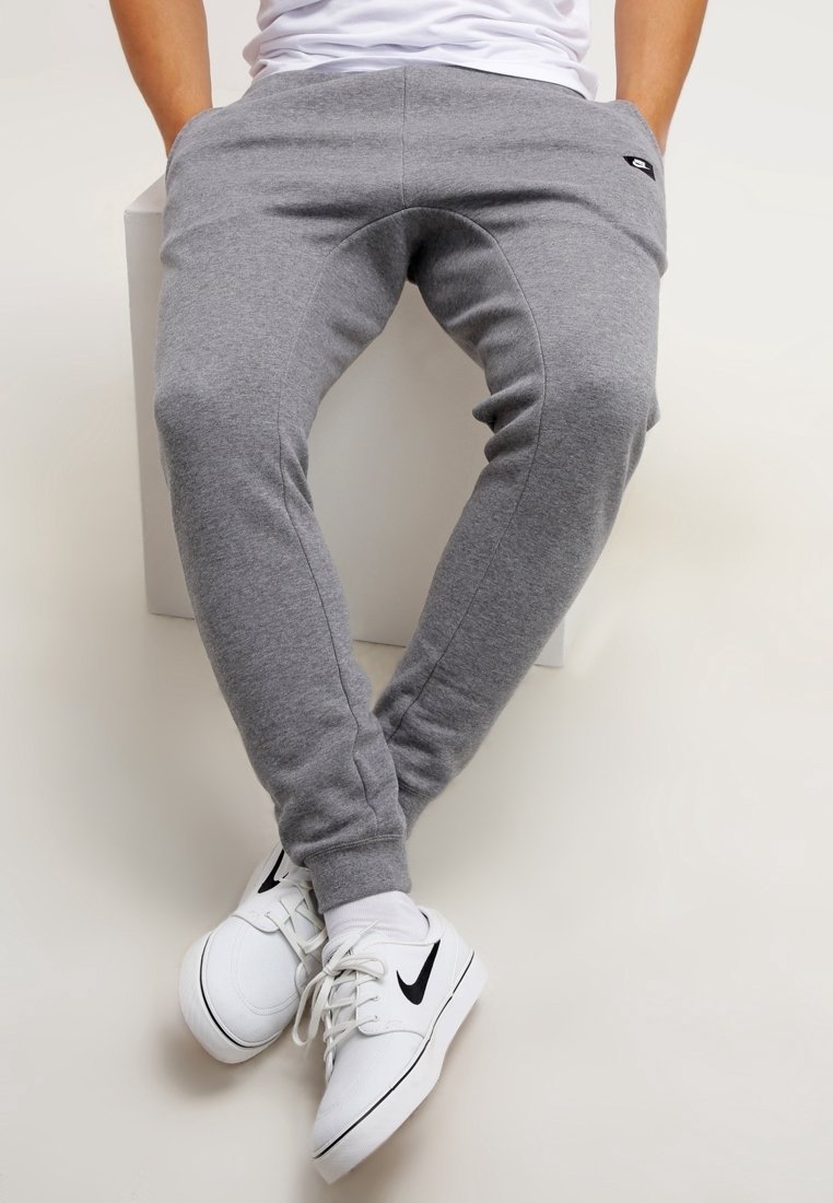Grey cotton sweatpants with a tapered design and elastic cuffs. Features a small black logo on the left thigh. Paired with white sneakers.