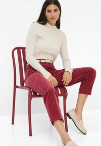 Geribbelde crèmekleurige coltrui, bordeaux corduroy broek met opgerolde boorden, beige loafers, zittend op een rode metalen stoel tegen een witte achtergrond.