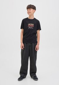 Crna pamučna majica s grafikom "JACK & JONES" u crvenoj i narančastoj boji. Opuštene tamne traperice i crne tenisice upotpunjuju odjevnu kombinaciju.