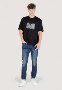 Un joven se encuentra de pie, de frente, con las manos en los bolsillos, vistiendo una camiseta gráfica negra, jeans azules y zapatillas blancas sobre un fondo liso.