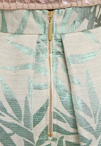 Un indumento aderente realizzato in tessuto beige chiaro con motivi a foglia verde, una cerniera in metallo dorato e dettagli plissettati vicino alla vita.