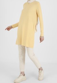 Henkilö, joka pukeutuu vaaleankeltaiseen pitkähihaiseen tunikaan, kermaisiin housuihin, vaaleabeigeihin lenkkareihin ja vaaleanbeigeeseen huiviin, valkoista taustaa vasten.