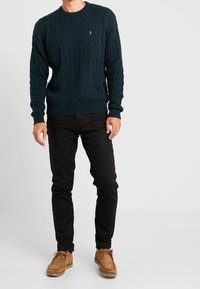 Donkergroen kabelgebreide trui met geribbelde manchetten en zoom, gecombineerd met zwarte slim-fit jeans en bruine suède veterlaarzen.