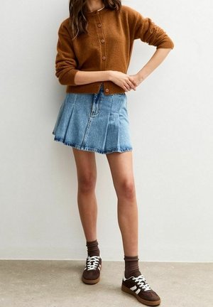 Femme portant un cardigan marron boutonné, une jupe en jean plissée bleue, des chaussettes marron et des baskets marron avec des lacets blancs, debout sur un sol en béton.