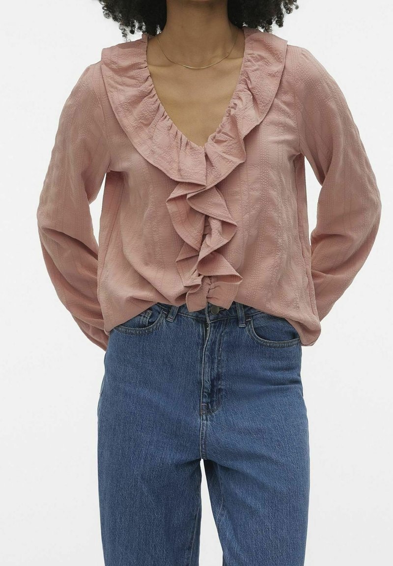 Blouse rose avec col à volants, manches longues et tissu texturé, associée à un jean bleu taille haute à coupe décontractée.