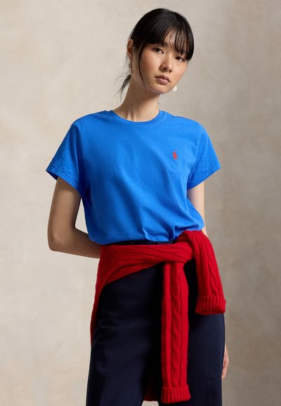 Frau trägt ein leuchtend blaues T-Shirt mit rotem Logo, marineblaue Hose und einen roten Zopfpullover um die Taille gebunden, steht vor einem einfarbigen Hintergrund.