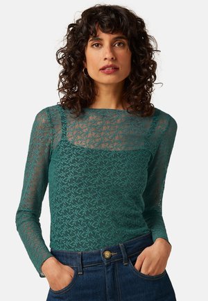 Kvinde med krøllet mørkt hår iført en langærmet teal blondebluse og blå jeans, står med hænder i lommerne mod en hvid baggrund.