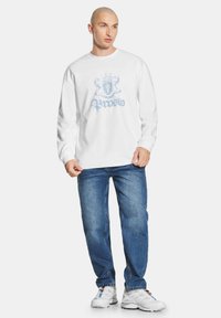 Biała koszulka z długim rękawem z jasnoniebieskim wzorem na przodzie, w połączeniu z niebieskimi dżinsami i białymi butami sportowymi.