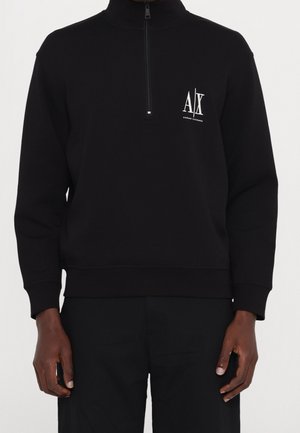 Pull-over noir à manches longues avec fermeture éclair quart de zip et logo Armani Exchange blanc sur la poitrine gauche, porté par une personne à la peau foncée.