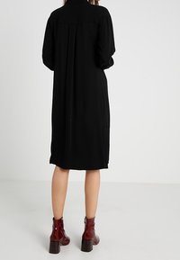 Abito camicia nero con maniche lunghe, vestibilità ampia, colletto abbottonato e dettagli plissettati sul retro, abbinato a stivaletti marroni alla caviglia.