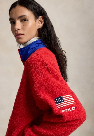 Γυναίκα με μακριά σκούρα μαλλιά, ντυμένη με κόκκινο φλις μπουφάν με μπλε γιακά και ένα κονκάρδα με την αμερικανική σημαία και την επιγραφή «POLO» στο μανίκι.