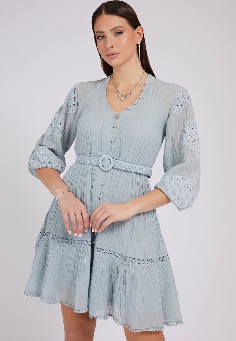 Lichtblauwe jurk van een gestructureerde stof, v-hals, knopen aan de voorkant en een gevlochten riem. Bevat kantaccenten op de mouwen en de zoom.