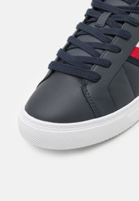 Sneaker in pelle blu con suola in gomma bianca, dotato di lacci piatti blu e striscia rossa di accento sul lato, texture liscia e design semplice.
