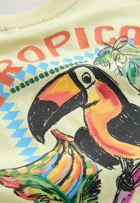 Tucan colorat cu cioc mare așezat lângă banane și frunze verzi, cu text roșu "TROPICAL" deasupra pe un fundal deschis de țesătură.