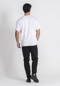 Weißes T-Shirt mit kurzen Ärmeln aus Baumwollstoff, kombiniert mit schwarzen Jeans und schwarzen Sneakers, von hinten an einem einfarbigen Hintergrund gesehen.