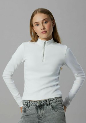 Weißes, geripptes Langarm-Oberteil mit Reißverschluss und hohem Kragen, kombiniert mit hellgrauen Jeans aus Denim, die Knopf- und Taschen-Details aufweisen.