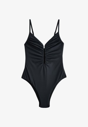 Zbliżenie osoby w czarnym bodysuit z wysokim wycięciem, pokazujące bok tułowia, biodro oraz górną część uda na neutralnym tle.