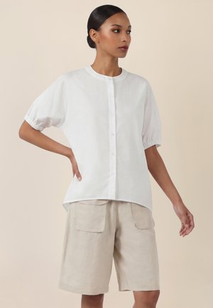 Frau trägt eine weiße, kurzärmelige Bluse mit Knöpfen und beige knielange Shorts mit aufgesetzten Taschen vorne, steht vor einem neutralen Hintergrund.