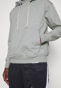 Personne portant un sweat à capuche Nike gris clair avec cordons, poche avant, et un short Nike noir avec des panneaux blancs sur les côtés.