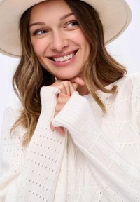 Młoda kobieta z jasnobrązowymi włosami uśmiecha się, nosząc biały sweter z dzianiny i beżowy kapelusz z szerokim rondem, z dłońmi przy brodzie.