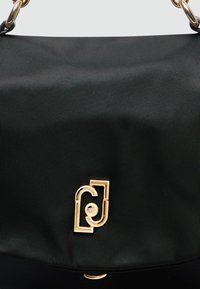 Crna najlon torba s teksturiranom površinom, koja se ističe istaknutim zlatnim kopčom u obliku logotipa, te ima strukturirani dizajn s preklopom.