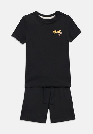 Μαύρο κοντομάνικο T-shirt με το κείμενο «PLAY» και κίτρινα αποτυπώματα πατούσας στο στήθος, σε συνδυασμό με μαύρα σορτς με κορδόνι.