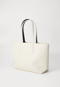 Sac fourre-tout blanc en matériau texturé, doté de deux longues poignées noires, avec une forme rectangulaire spacieuse et un design minimaliste.