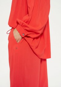 Koraalkleurige blouse met lange mouwen en gathered cuffs, voorzien van een zak en knoopdetail aan de zijkant. Soepele, vloeiende stoftextuur.