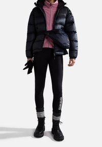 Zwarte pufferjack met capuchon, roze rits-innerlaag, zwarte legging, grijze beenwarmers en zwarte veterschoenen met dikke zolen.