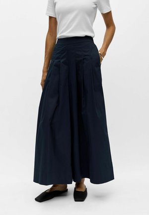Femme portant un haut blanc à manches courtes avec une jupe bleu marine taille haute, jambes larges et chaussures plates noires, debout avec une main dans la poche.