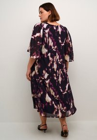 Robe maxi violet foncé avec des motifs abstraits crème et bordeaux ; tissu léger, coupe ample et manches volantées. Sandales à talons noires avec lanières.