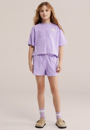 Ragazza giovane in piedi che indossa una maglietta a maniche corte color lavanda e pantaloncini abbinati con bordi smerlati, calzini a righe e scarpe beige slip-on.