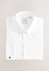 Chemise blanche à manches longues pliée, col à boutons, un seul bouton de manchette noir sur le poignet gauche et étiquette "NEXT TAILORING" à l'intérieur du col.