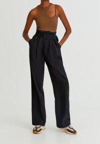 Personne portant un haut sans manches marron avec découpe, un pantalon large taille haute noir ceinturé, et des baskets blanches avec des accents verts et violets.