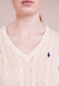 V-ringad stickad tröja i krämfärg, med kabelstickat mönster och ribbade kanter, samt en liten marinblå logotyp på vänster bröst.