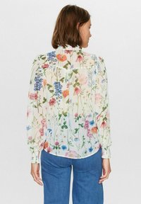 Blommig blus med långa ärmar, vit genomskinlig tyg, med en flerfärgad blommönster och rynkade manschetter. Rundad nederkant baktill.