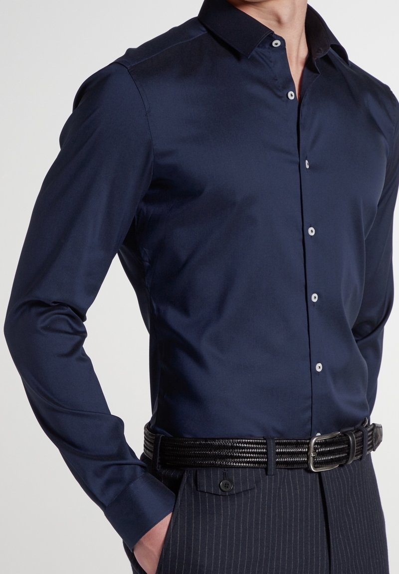 Homme portant une chemise ajustée bleu foncé rentrée dans un pantalon à rayures fines avec une ceinture tressée noire, main dans la poche, sur un fond uni.