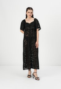 Black lace maxi dress with short sleeves, featuring a scalloped neckline and button detailing. Paired with black high-heeled sandals with bow accents.
