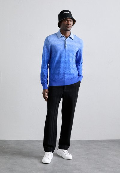 Polo s modrým gradientem a zigzagovým vzorem, kombinovaný s černými kalhotami v houndstooth vzoru a bílými teniskami. Černý klobouk ve tvaru kbelíku s logem.