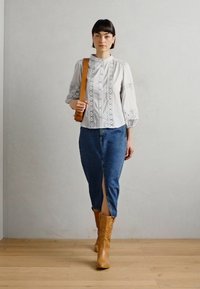 Balta blūze ar mežģīņu detalizāciju, apvienota ar zilu džinsa svārku ar priekšējo spraugu. Augsti brūni ādas zābaki papildina apģērbu.